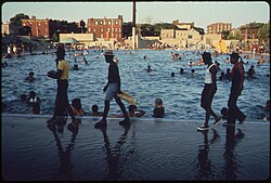 A 1974 color photograph of a public pool in Brooklyn being used by a large number of black people, with four people walking by in the foreground.